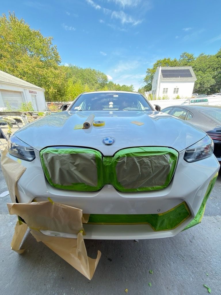 White BMW front with green painter’s tape and cardboard masking in a driveway, ready for paint.