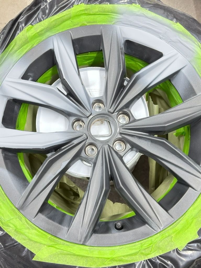 Car wheel rim with silver spokes, surrounded by bright green masking tape, in a repair or paint-prep setting