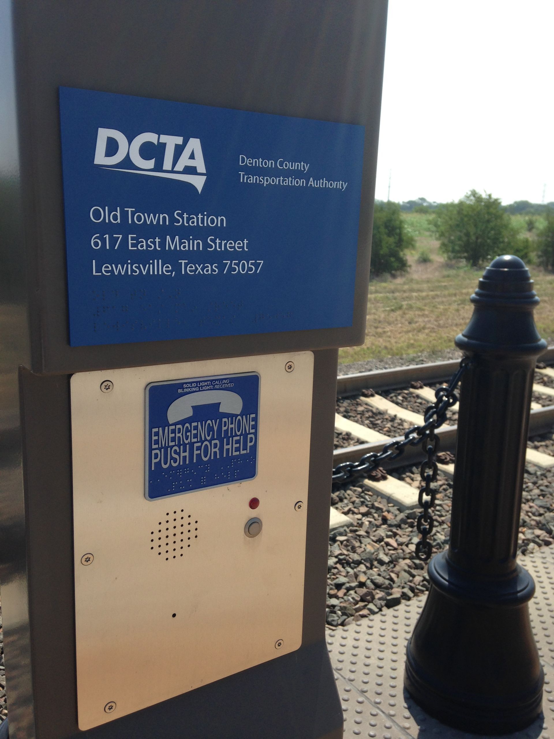 Blue DCTA transit sign beside tracks with emergency call box and black post near a rail line