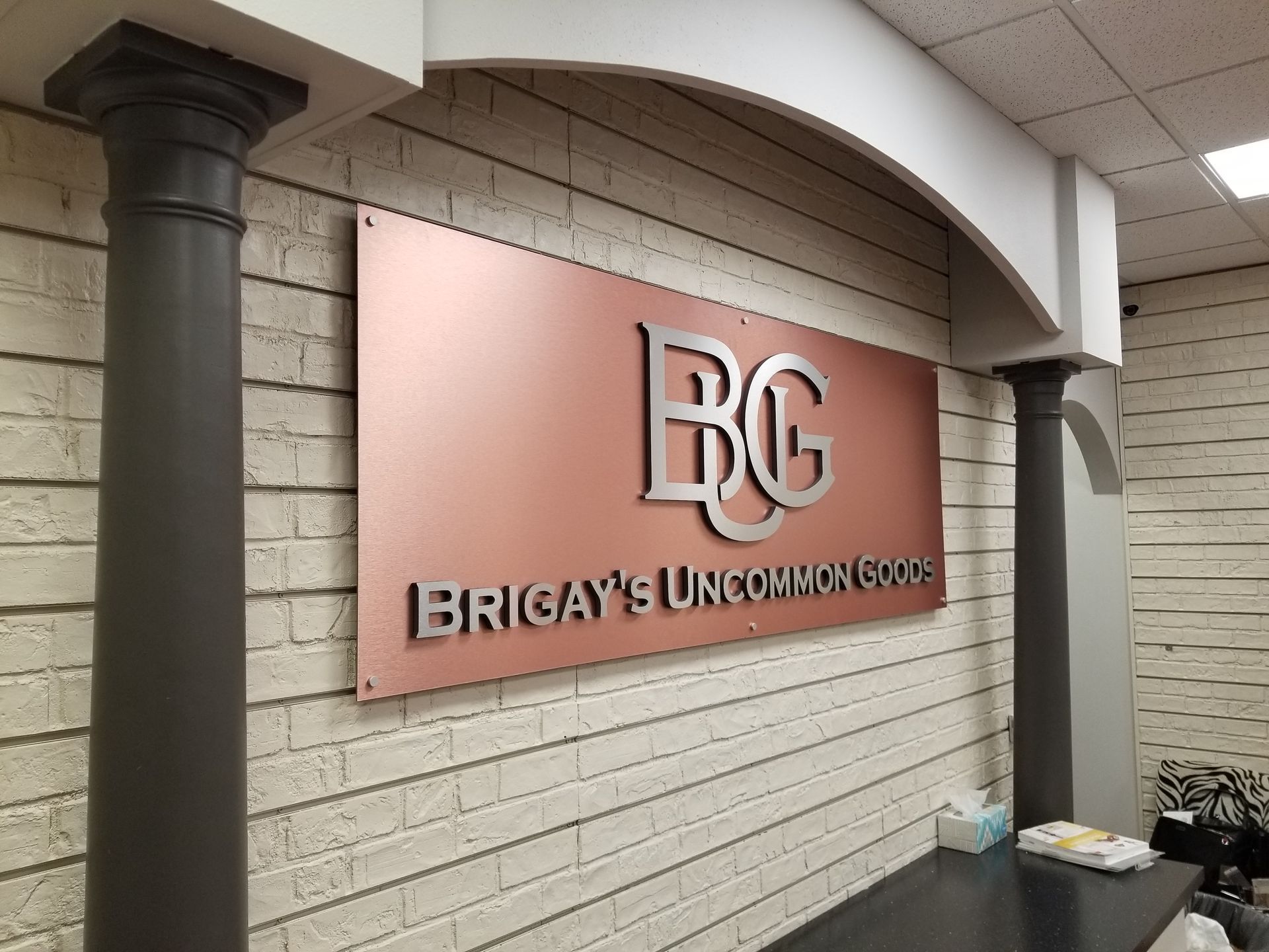 Bronze wall sign reading “BG” and “Brigly’s Uncommon…” in a hallway with white brick walls