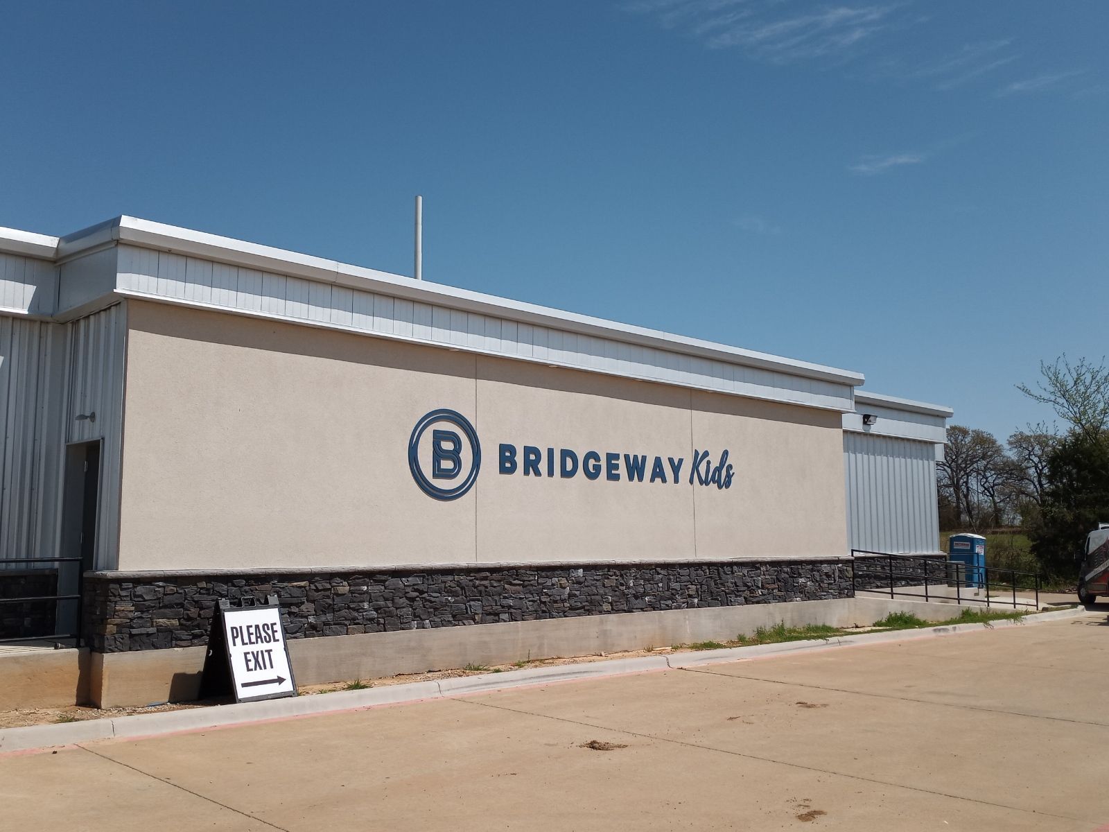 Bridgeway Club building with logo on beige wall and small sign in front under a clear blue sky