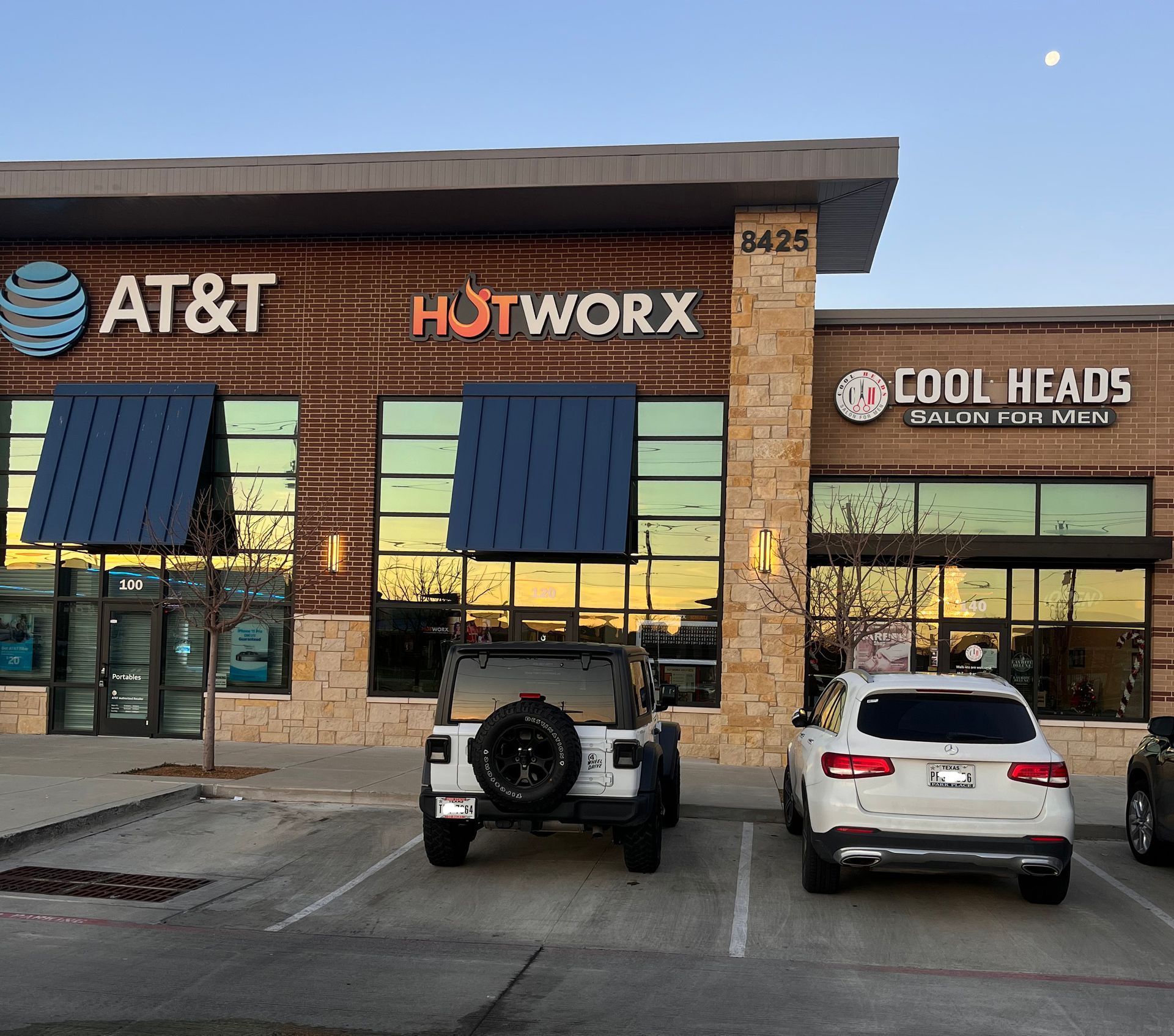 Strip mall storefronts for AT&T, Hotworx, and Cool Heads with parked cars at dusk