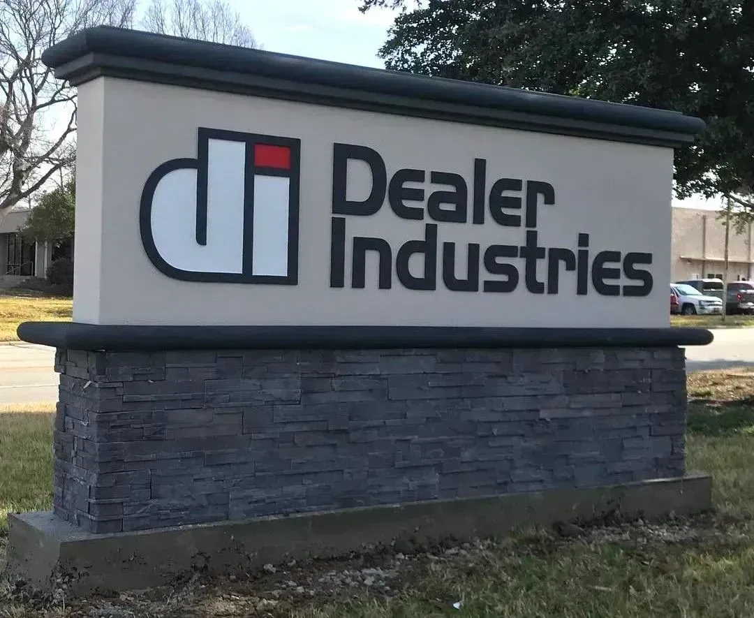 Dealer Industries sign on a brick base outdoors with black lettering and a red-and-white logo.
