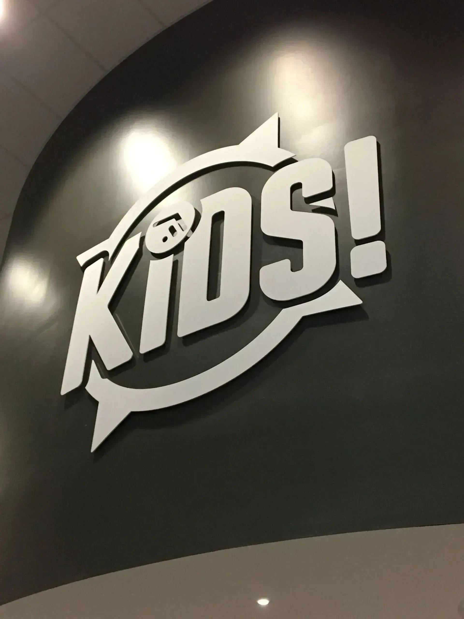 White “KIDS!” sign on a dark curved wall with a speech-bubble shape and spotlight reflection.