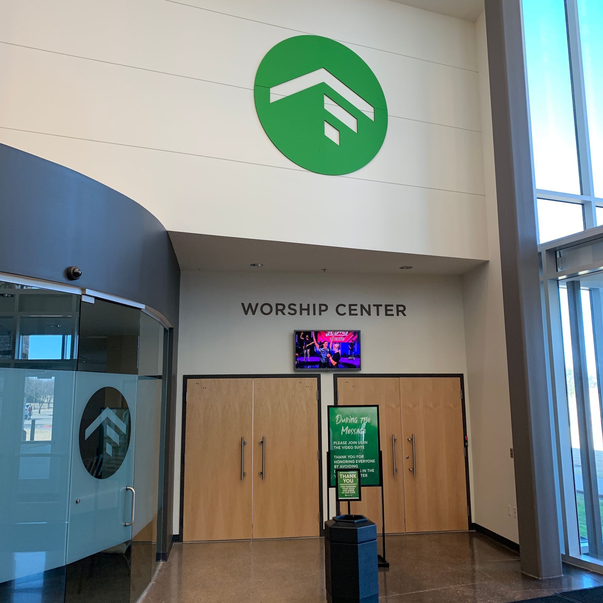 Worship center entrance with wooden doors, green logo, and a sign stand in a bright lobby