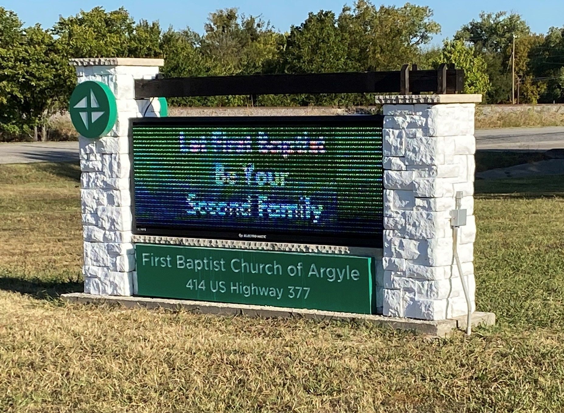 Church marquee sign in front of stone pillars on a sunny roadside lawn.