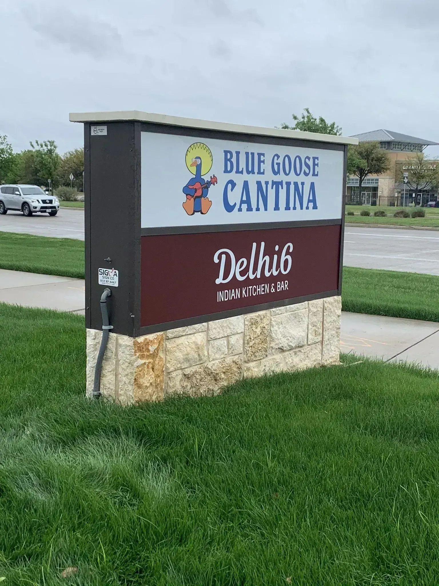 Blue Goose Cantina sign with Del Hib text on a stone base by a roadside grassy area