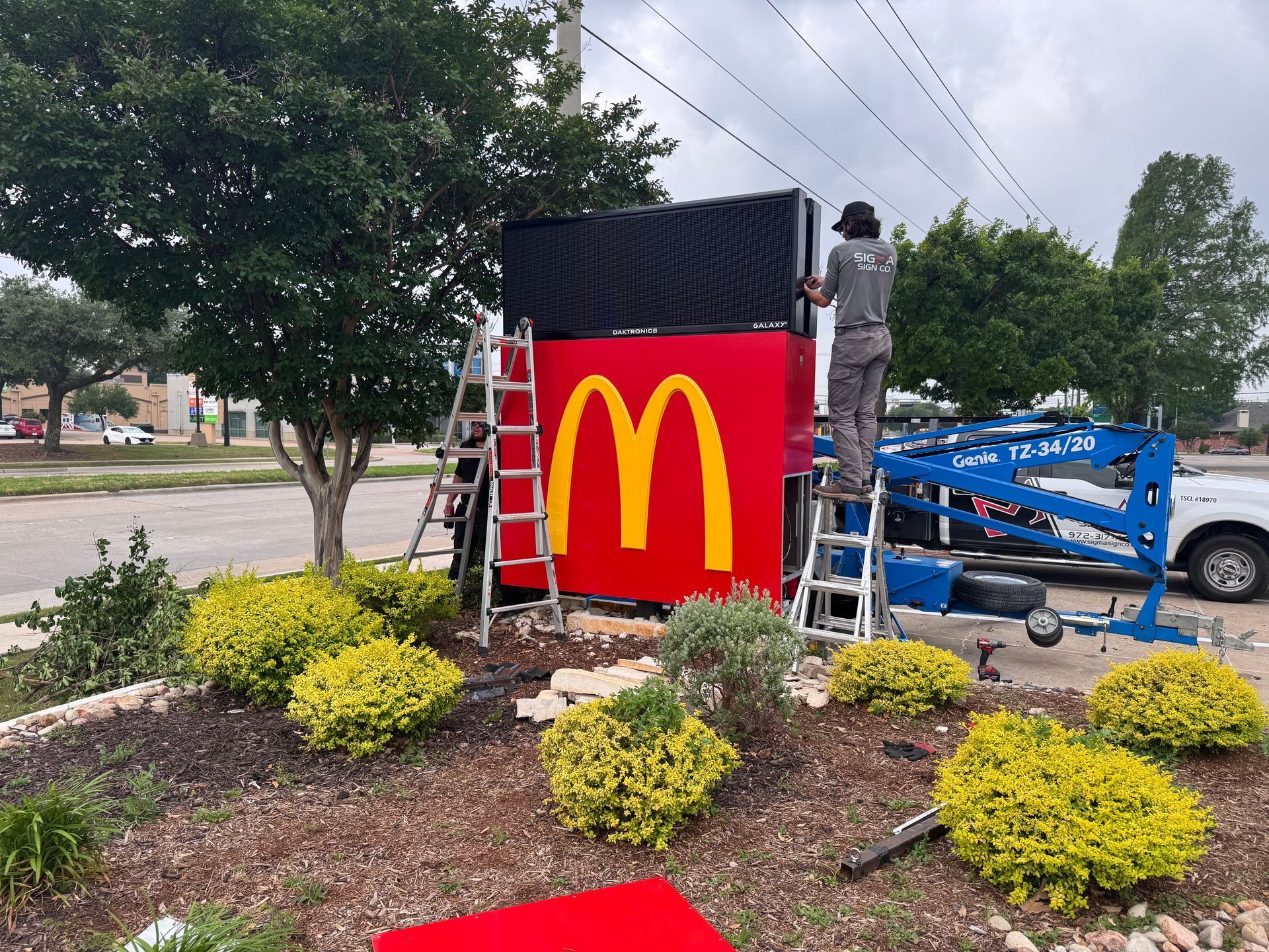 Worker installs McDonald’s sign beside a blue lift in a landscaped roadside area
