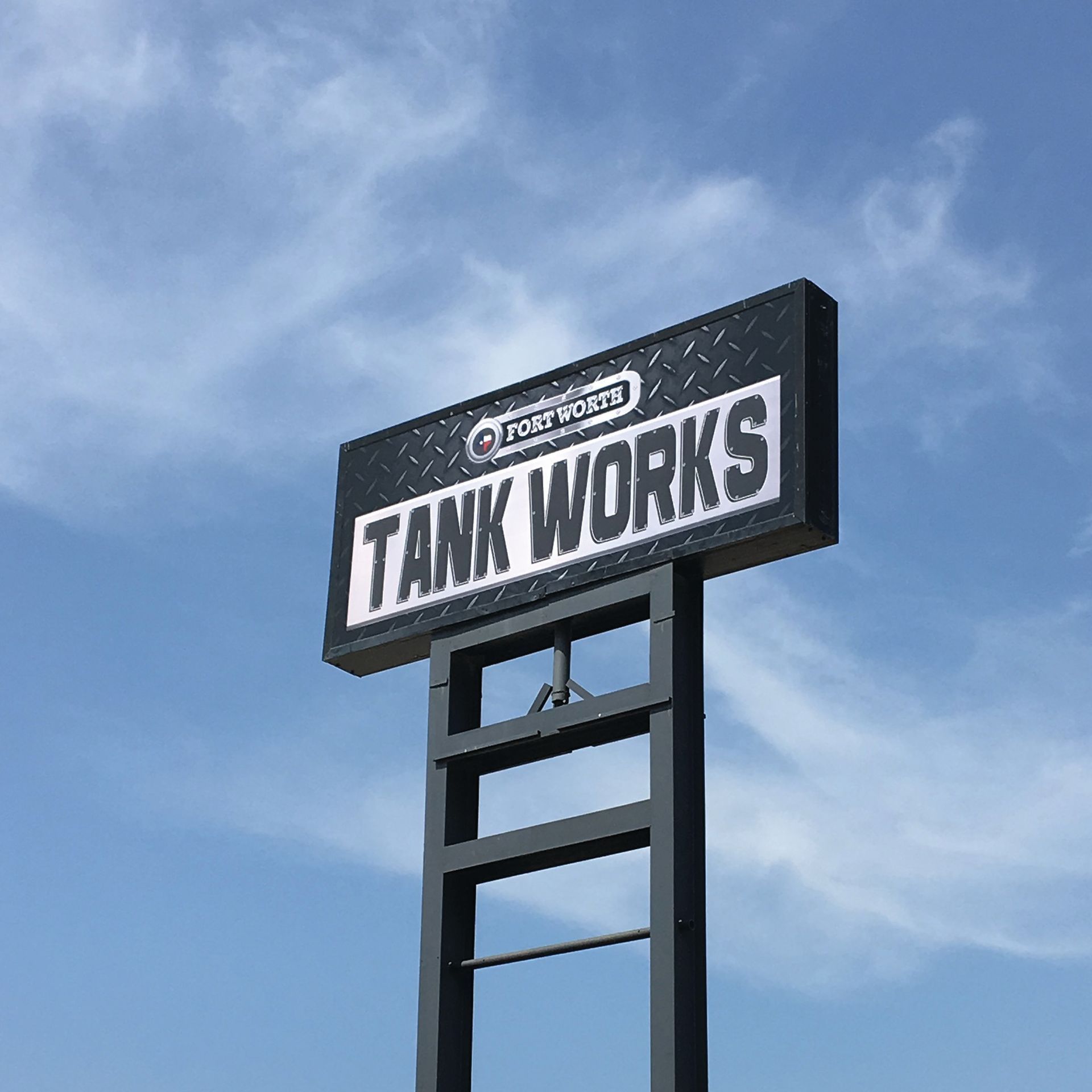 Black sign reading “TANK WORKS” on a tall pole against a blue sky
