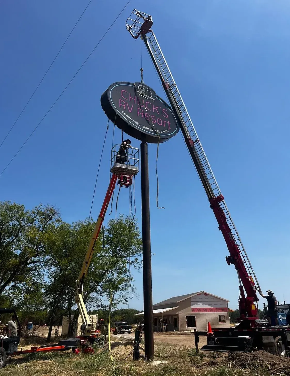 Two crane trucks hoist a large round sign on a tall pole against a clear blue sky.