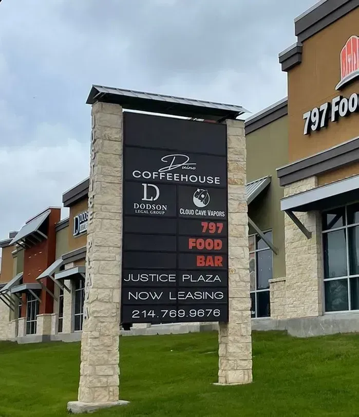 Coffeehouse plaza sign in front of storefronts on a grassy strip under a cloudy sky.