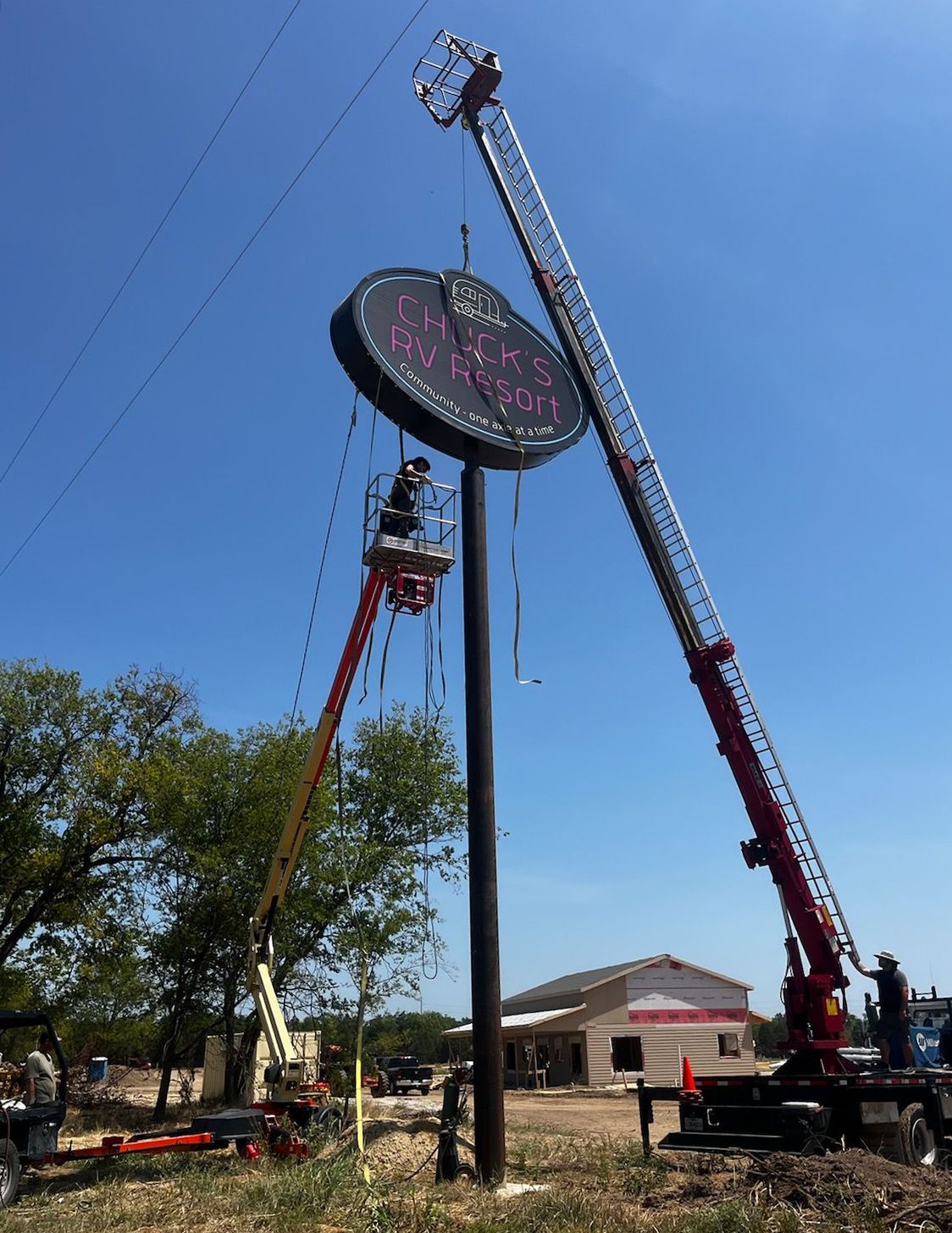 Two aerial lifts servicing a round sign. One worker in a lift basket is working on the sign. Blue sky background.