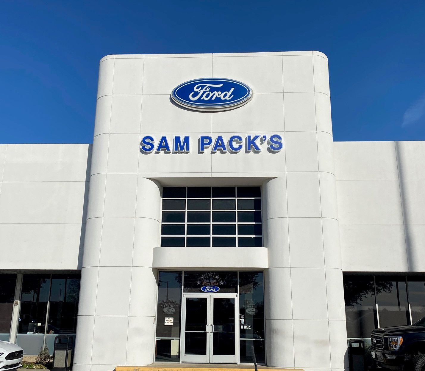 Ford dealership, Sam Pack's, front entrance with Ford logo and blue lettering on a white building, under a blue sky.