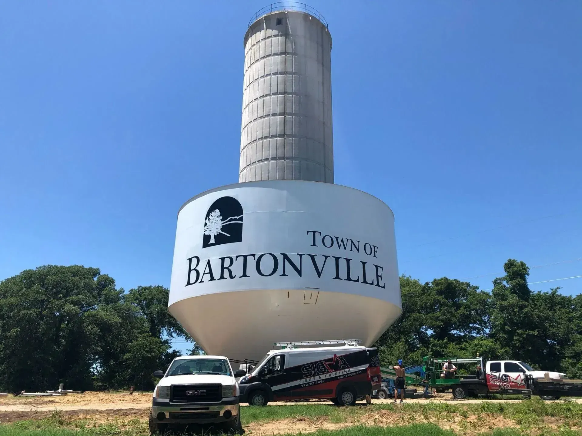 Water tower in Bartonville with Town of Bartonville logo; vehicles and trees in foreground under a blue sky.