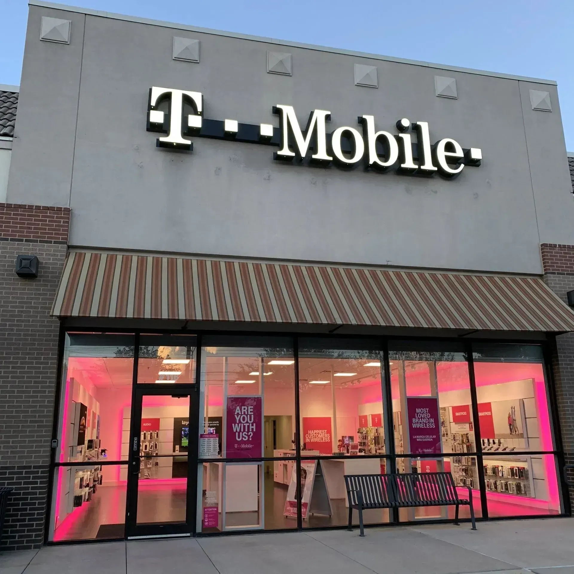 T-Mobile store exterior with illuminated pink interior and logo, awning, windows, and bench.