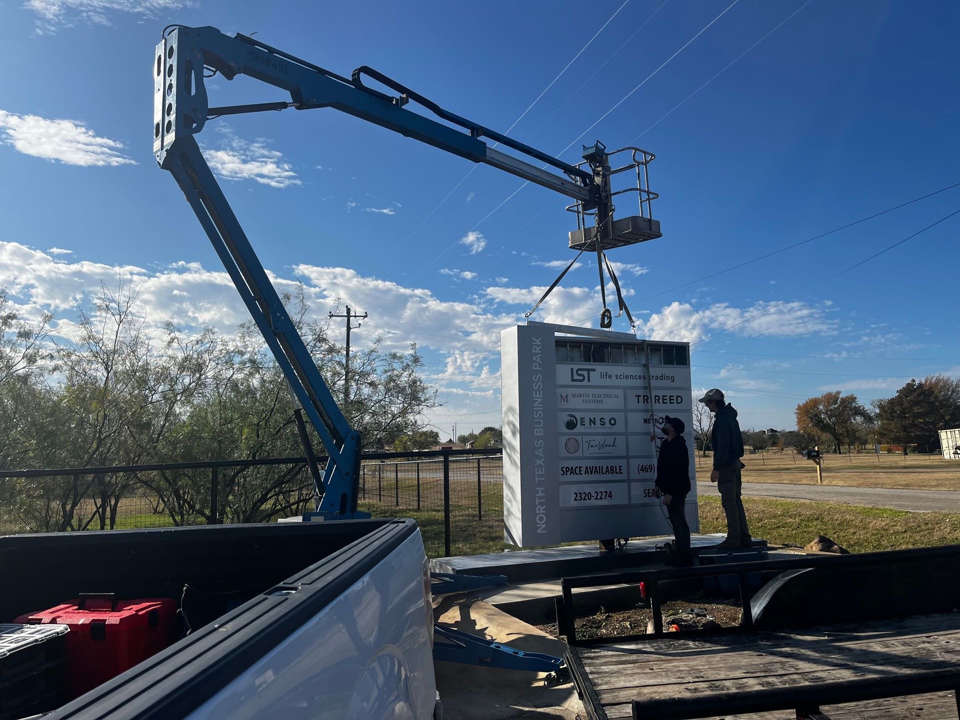 Workers operating a boom lift beside a large white panel outdoors under a blue sky