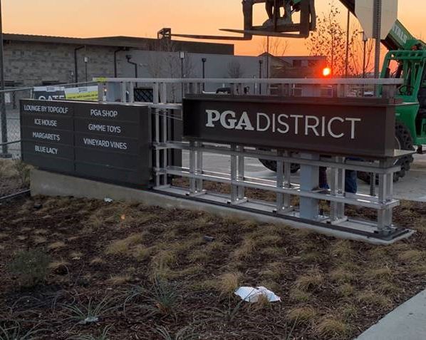PGA District sign at sunset beside a golf course entrance, with metal railings and dry grass.
