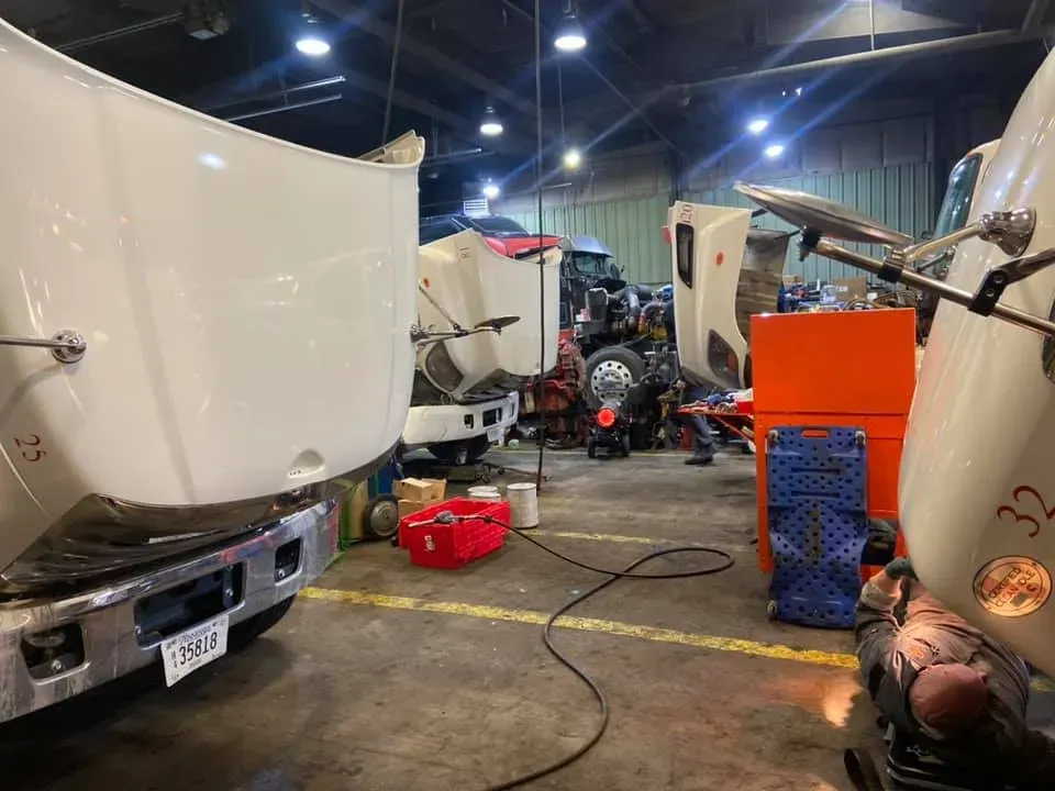 Trucks being repaired in a garage; hoods up, tools and equipment scattered about, worker visible in the background.