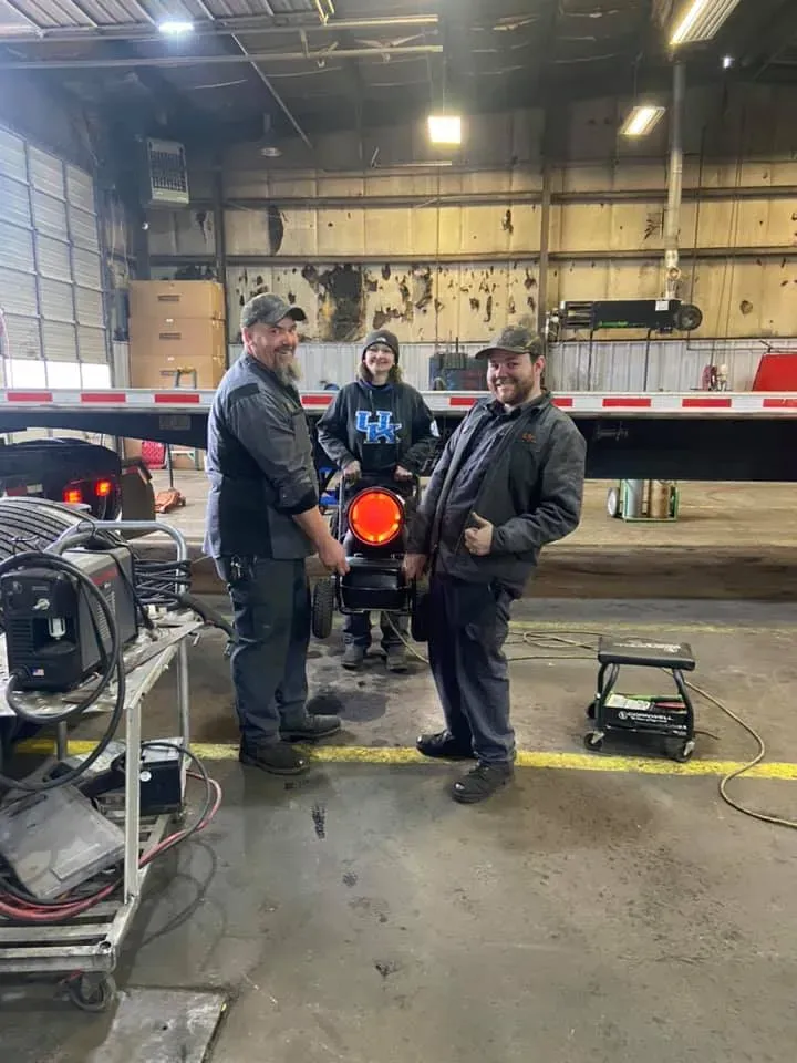 Three people stand near a truck in a garage. Two are smiling and holding a tail light, the other is in the middle.
