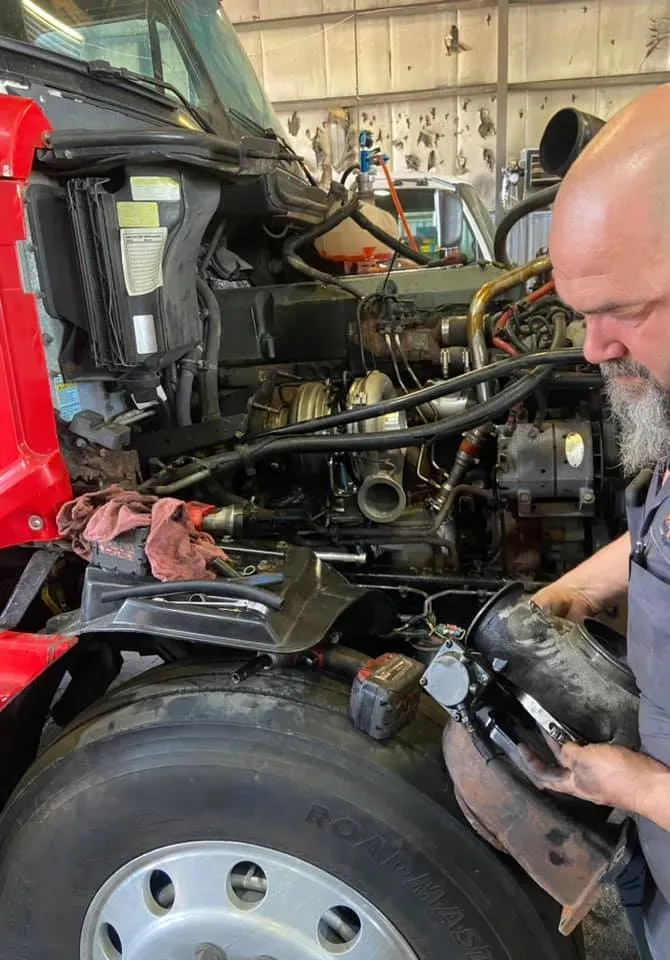 Mechanic working on a truck engine, removing a part. Red truck, gray-haired man in shop.