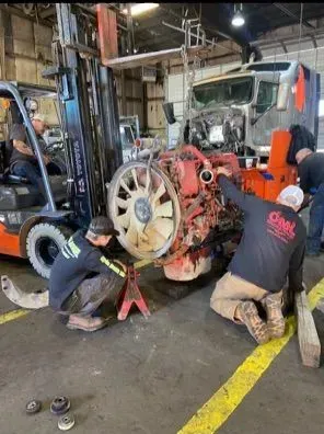 Workers servicing a truck engine in a garage; using a forklift and jack stands.