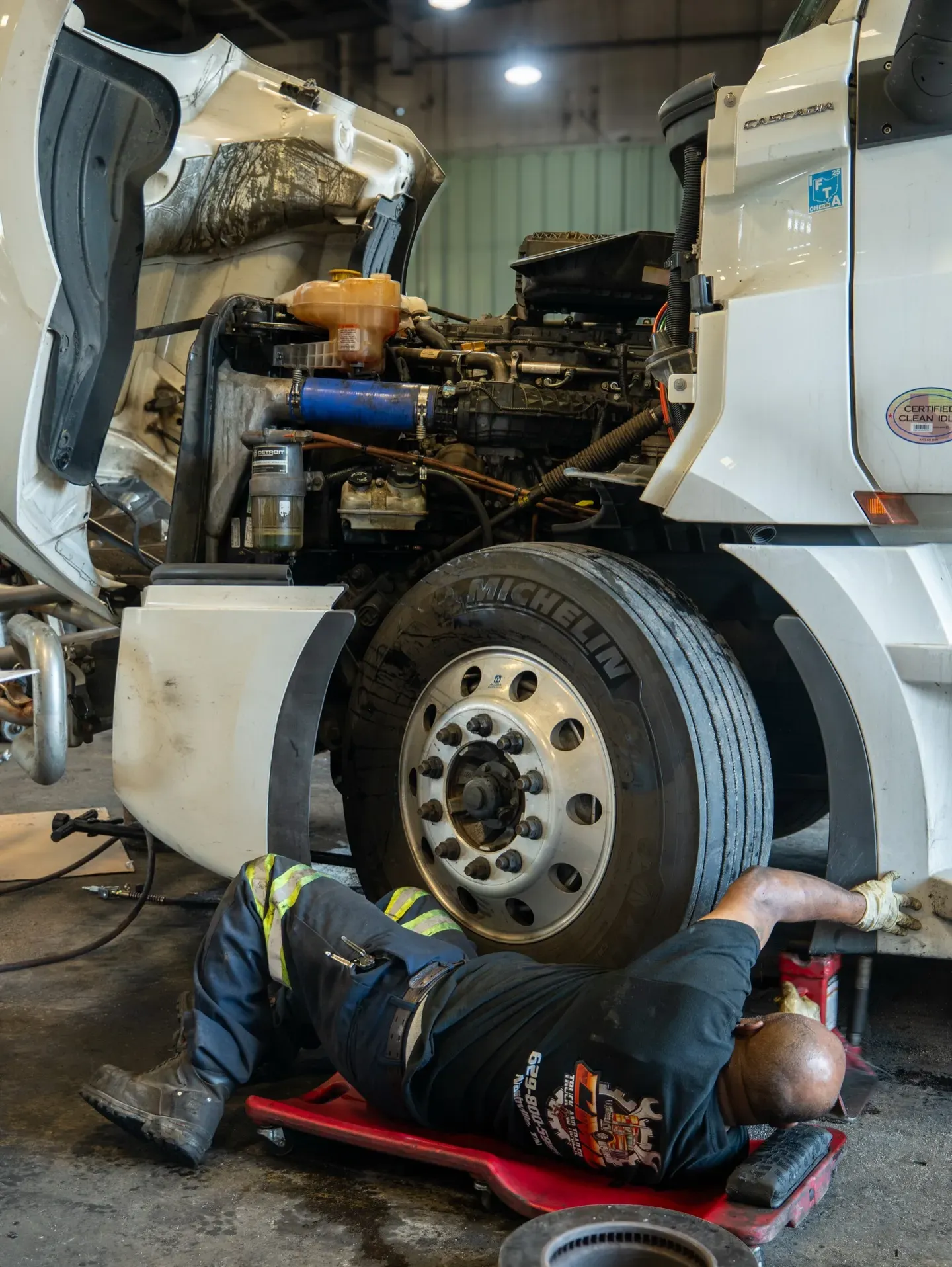 Mechanic working on a truck engine, lying on a creeper. The truck is white, in a garage setting.