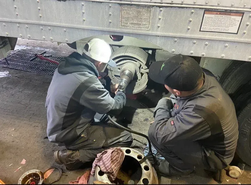 Two mechanics repair a truck axle under the vehicle. One uses a tool, the other observes.