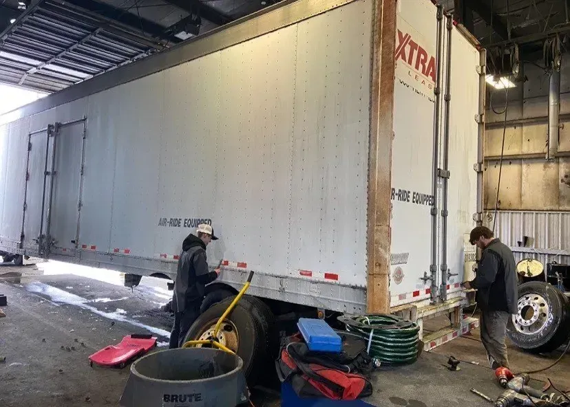 Two people working on a white XTRA trailer in a garage. Tools and tires are visible.