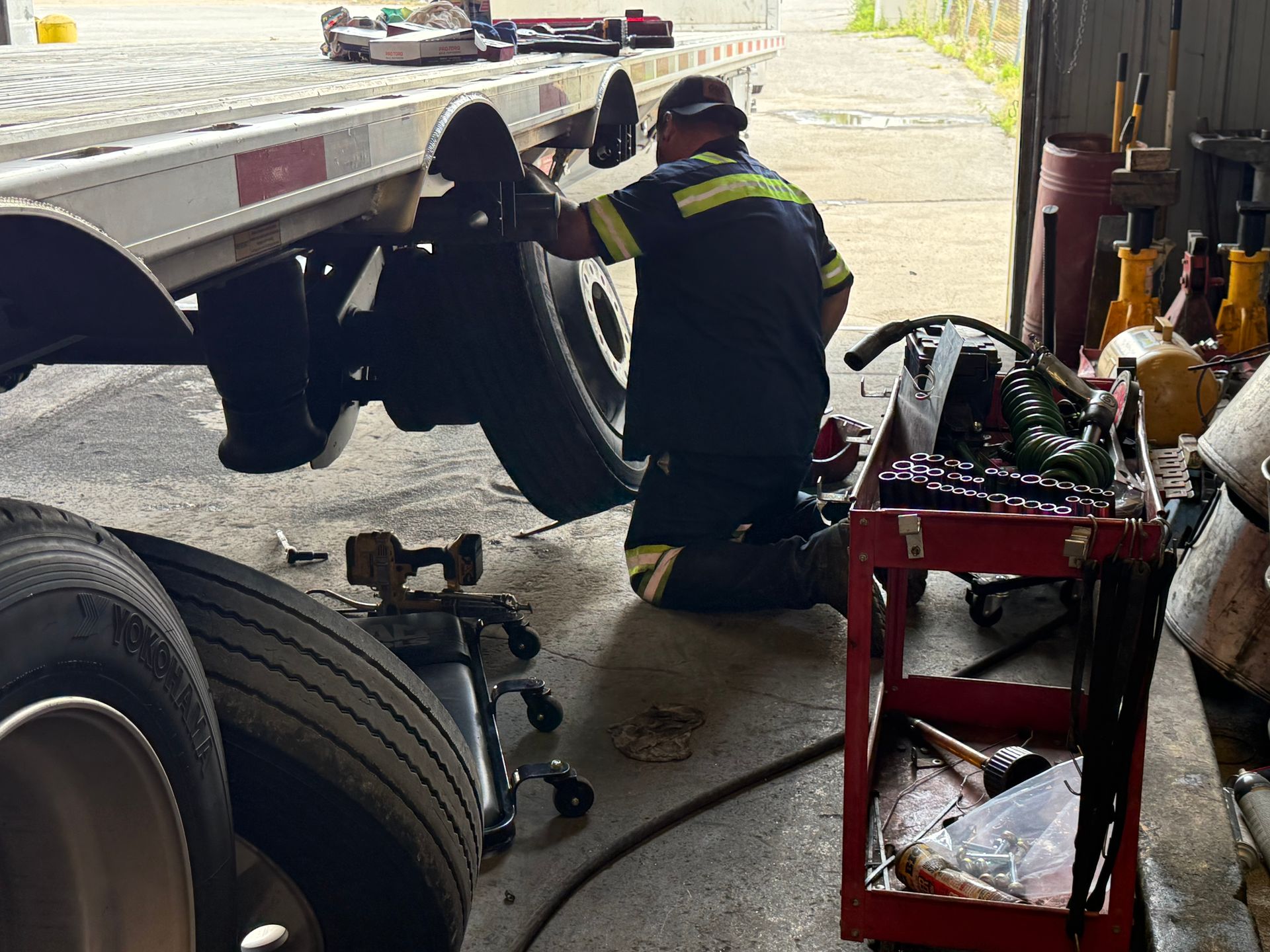 A technician in a high-visibility uniform kneels in a garage, repairing the wheel of a large flatbed trailer.