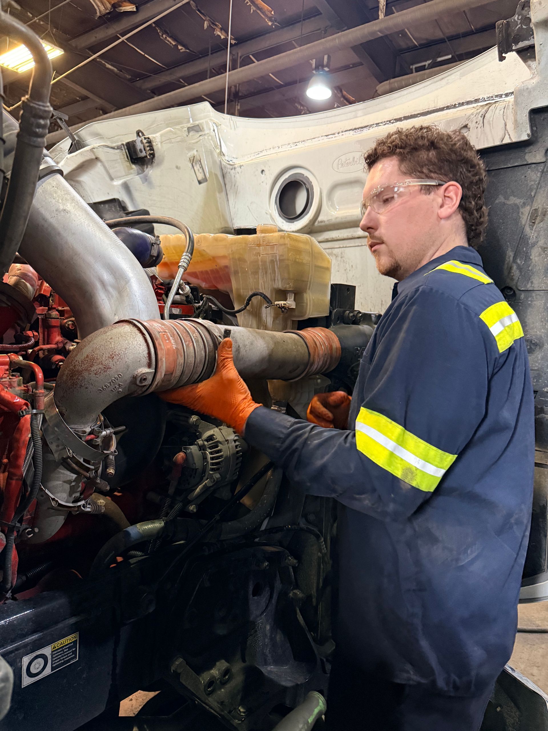 A person in a blue uniform and safety glasses works on the engine of a large truck in a workshop.