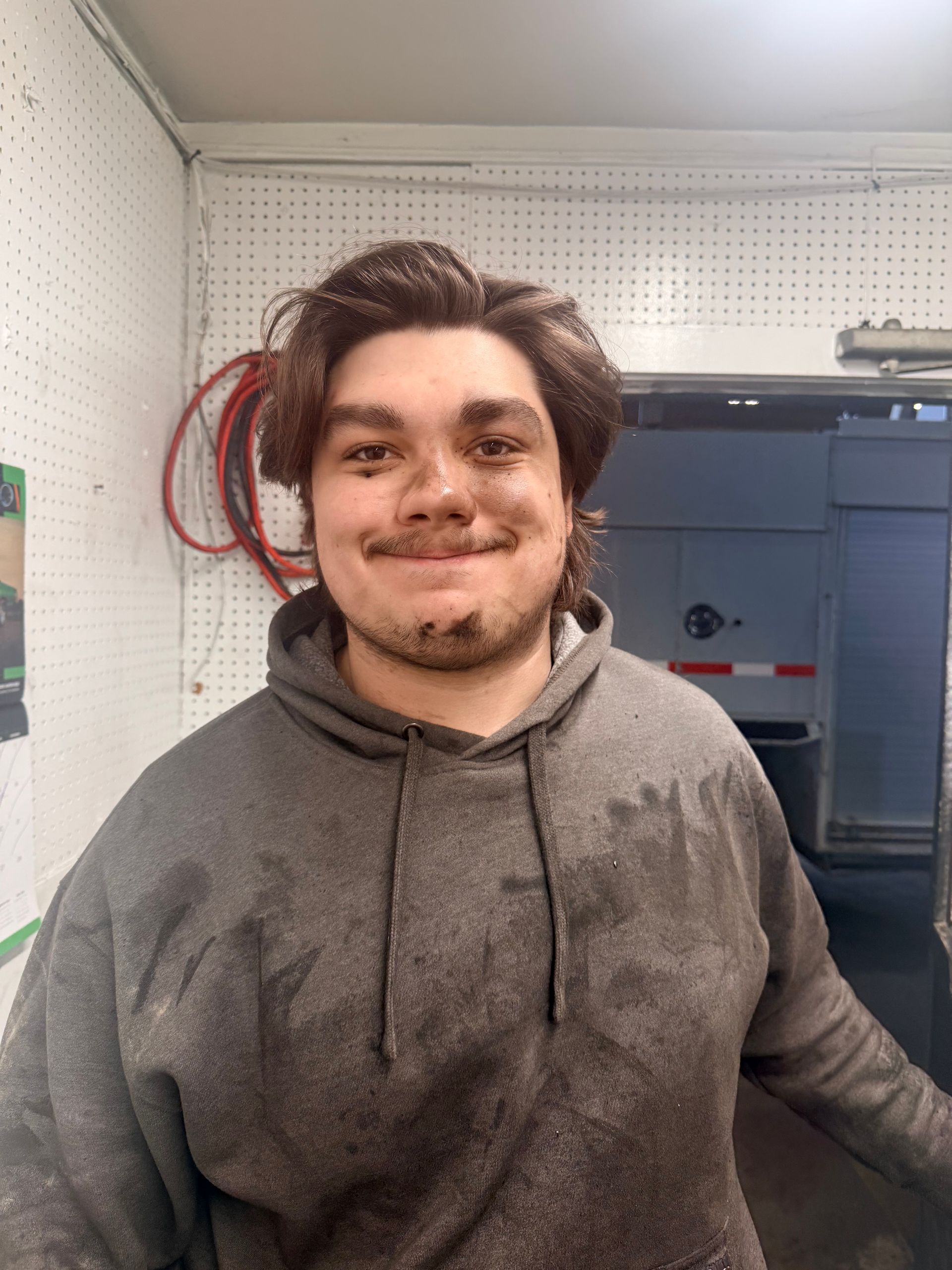 A person with messy hair and smudge marks on their face and gray hoodie stands in a workshop with a trailer behind them.