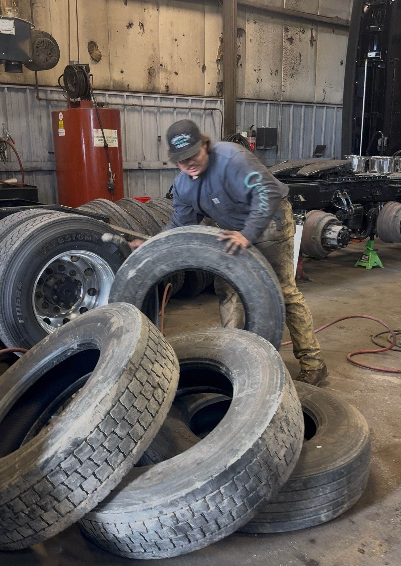 A mechanic in a garage setting rolls a large truck tire toward a stack of tires.