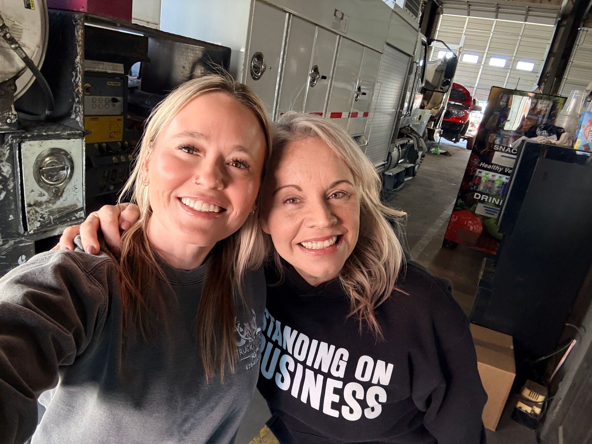 Two people posing for a selfie in a warehouse or garage setting, both smiling at the camera.