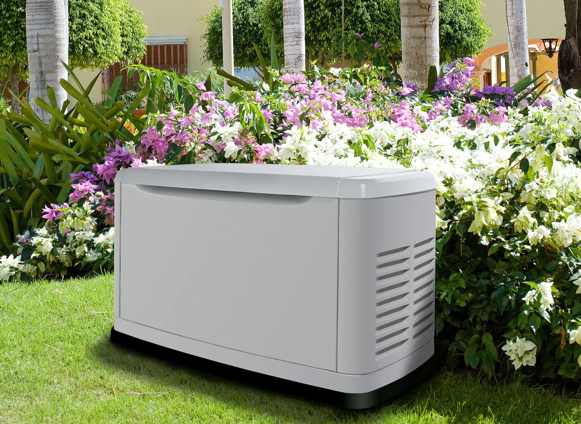 A gray generator in a garden, surrounded by plants and flowers.