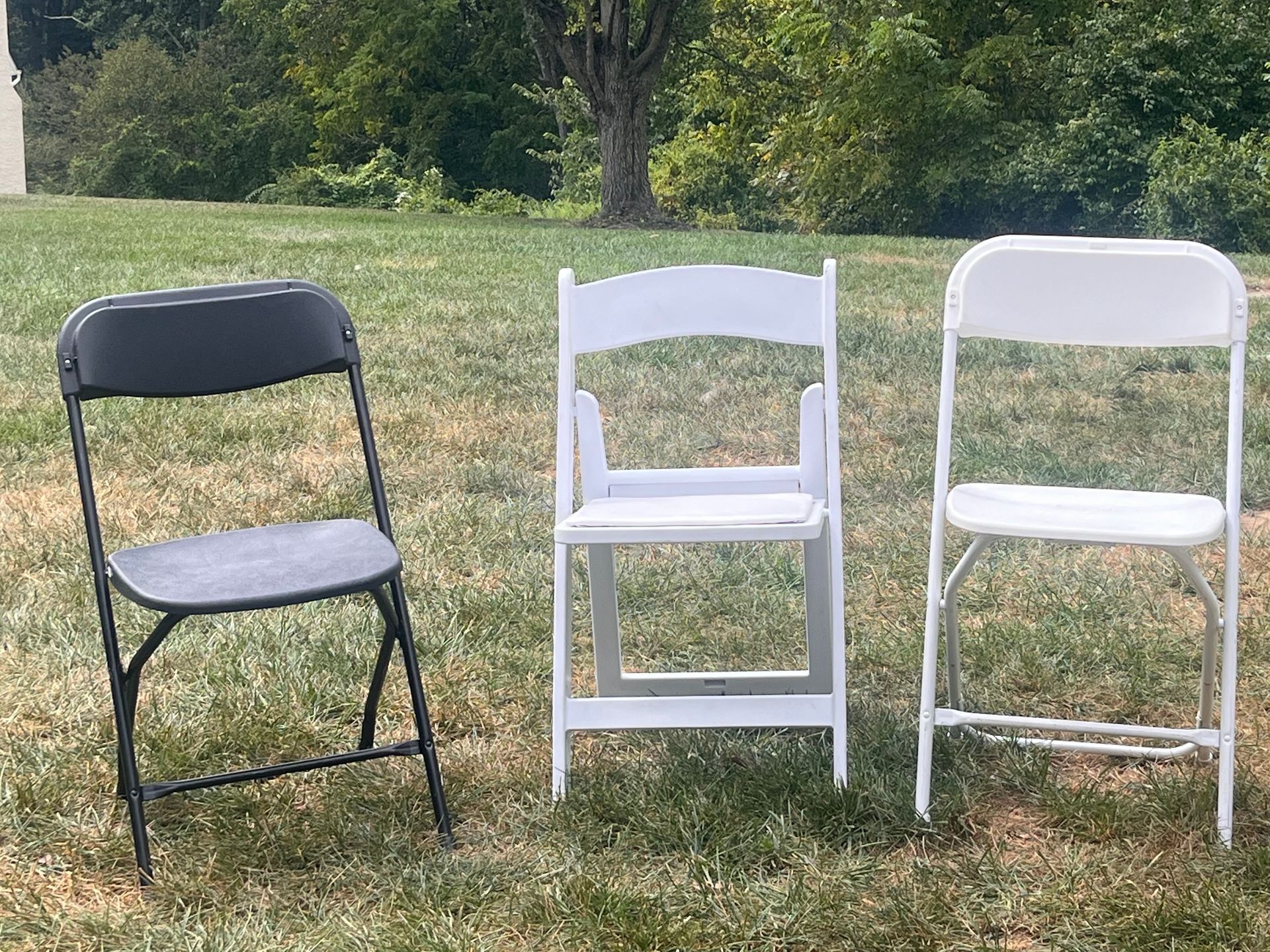 Three folding chairs in a grassy yard: black, white, and a white one with a decorative back, on a sunny day.