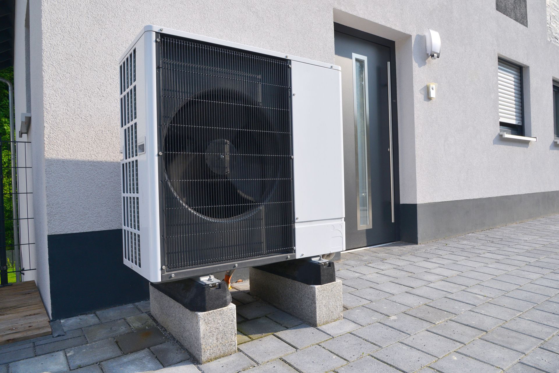 Air-source heat pump mounted on concrete blocks outside a white building with gray trim and a front door.