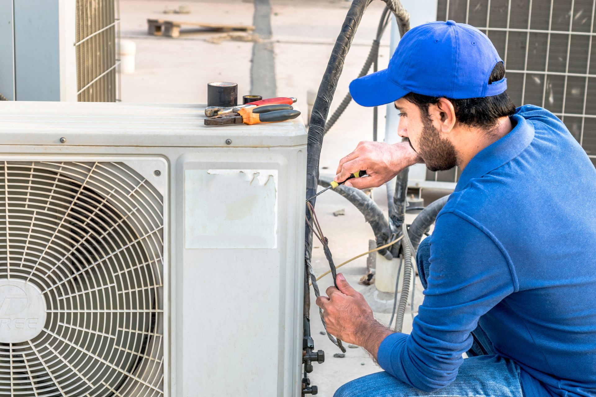 HVAC technician in blue works on rooftop AC unit, using tools.