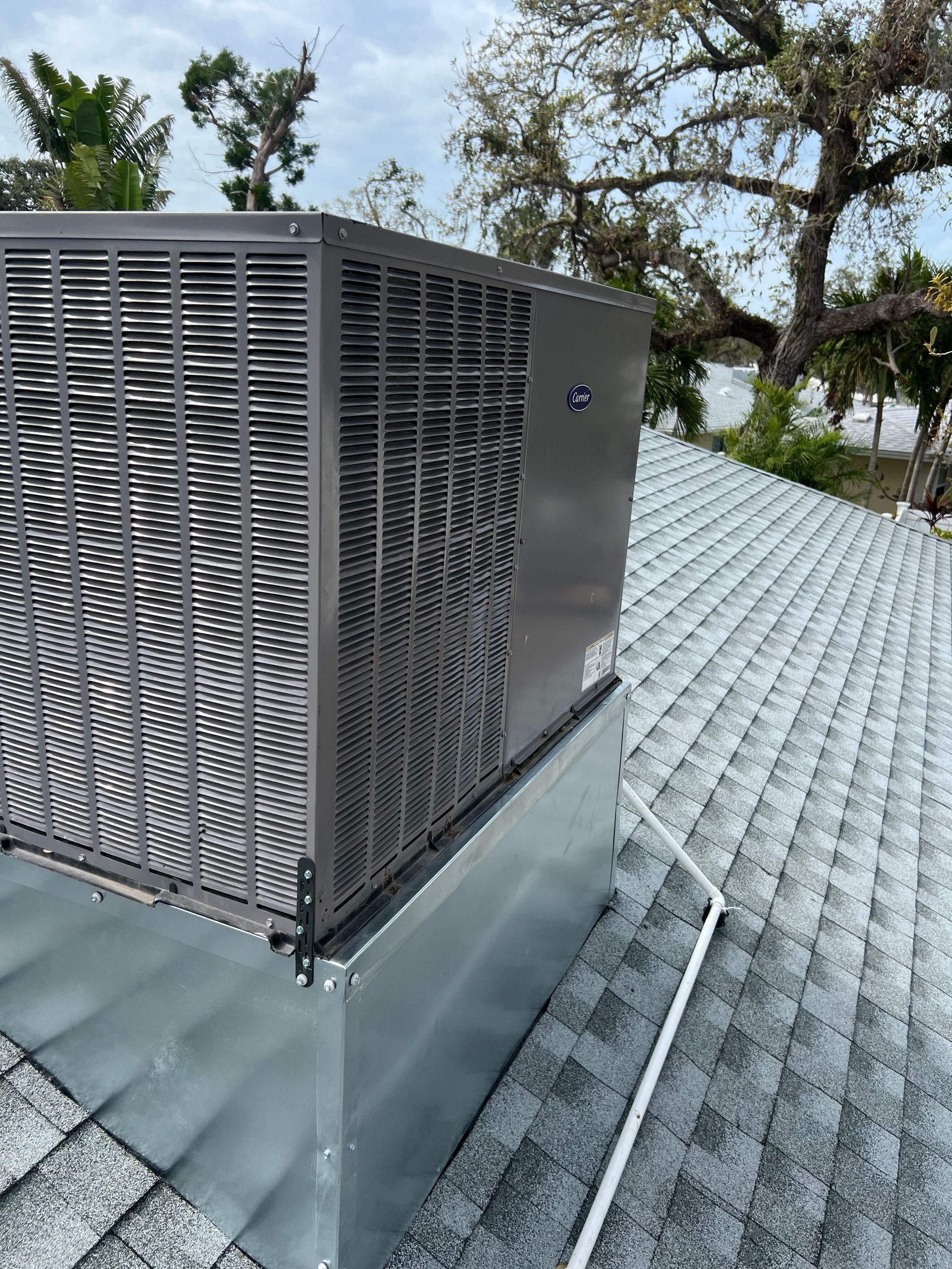An air conditioning unit sits atop a gray shingled roof, against a backdrop of trees and cloudy sky.