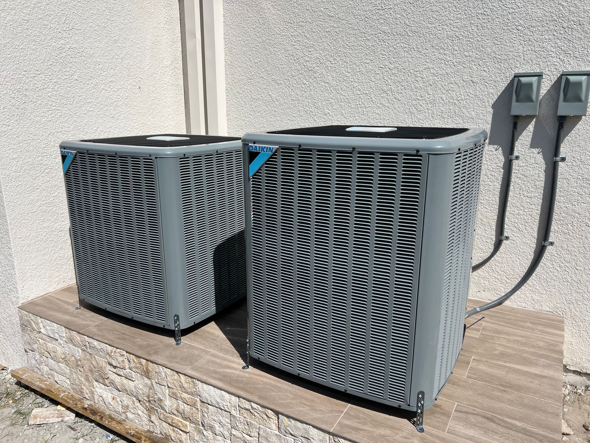 Two gray air conditioning units outside on a stone ledge, next to electrical boxes.