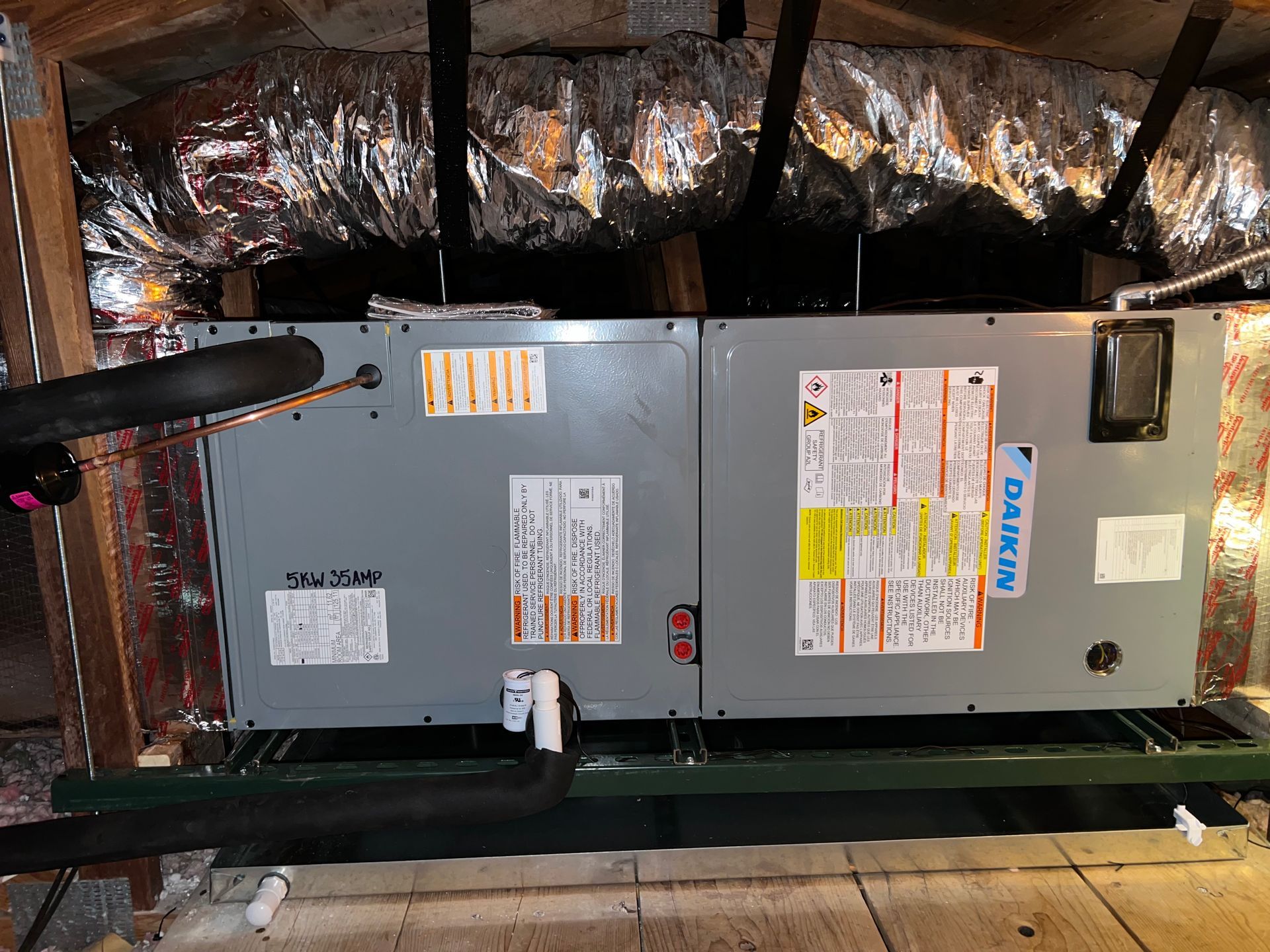HVAC unit in an attic. Gray metal box, Daikin brand, with ductwork and insulation overhead.