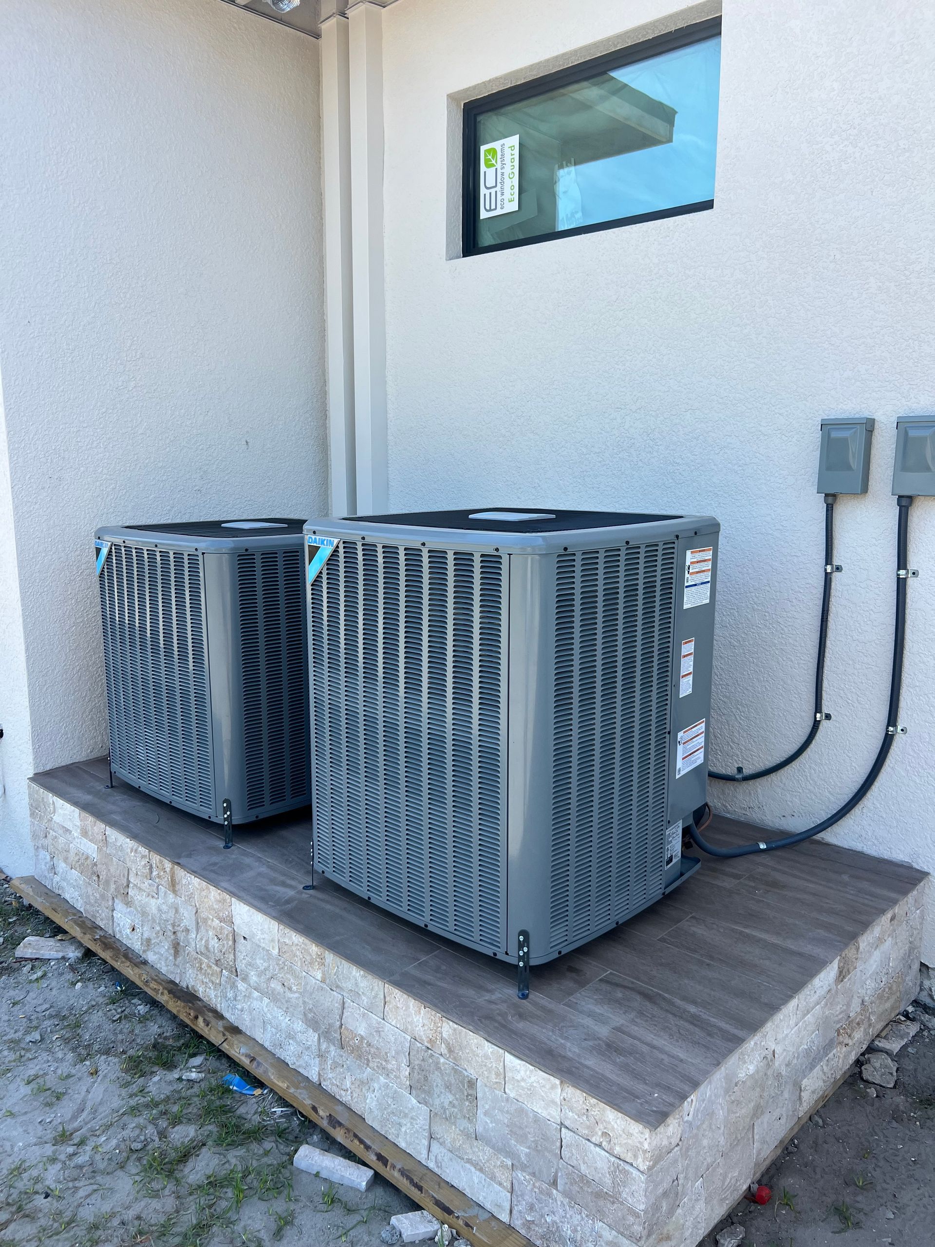 Two gray air conditioning units on a stone platform next to a white building with a window.