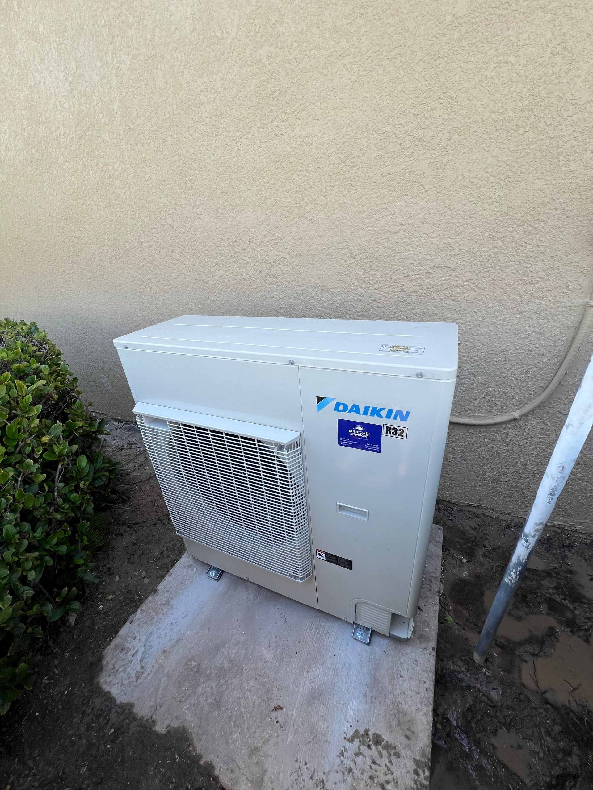 A Daikin air conditioning unit against a beige stucco wall and a small bush on a concrete pad.