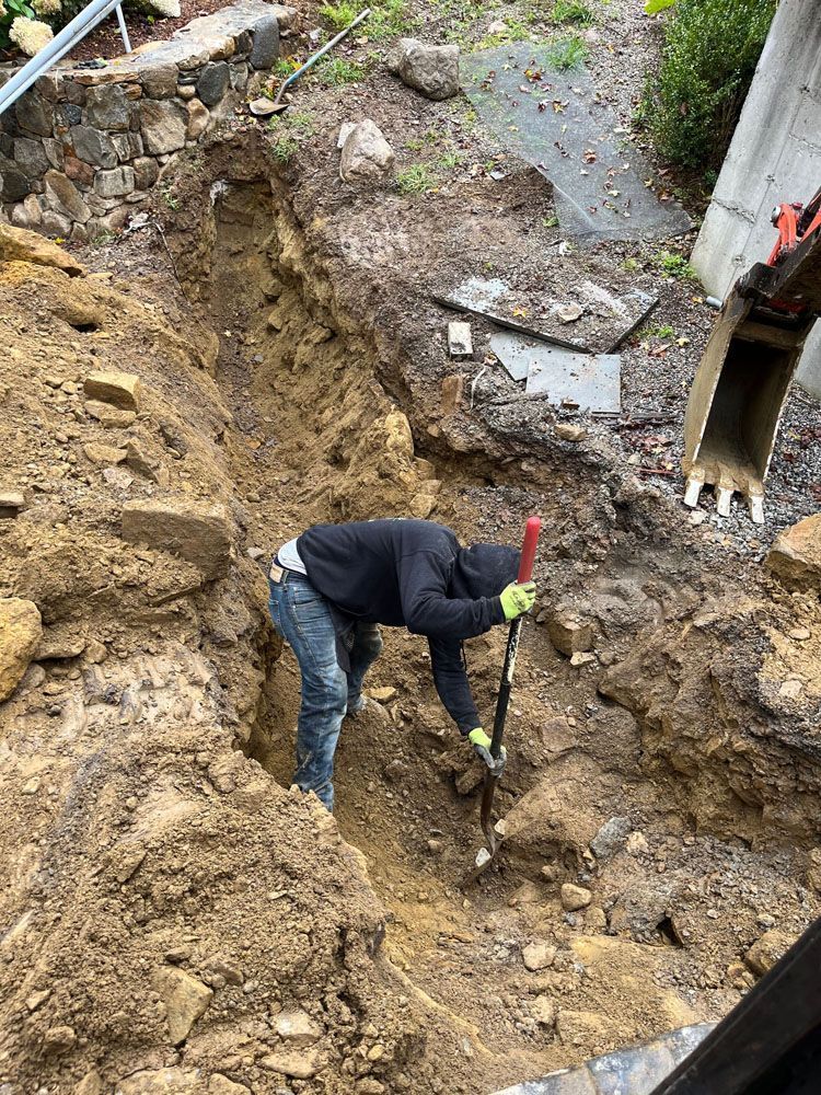 Person digging in a muddy landslide area beside a damaged structure with exposed pipes.