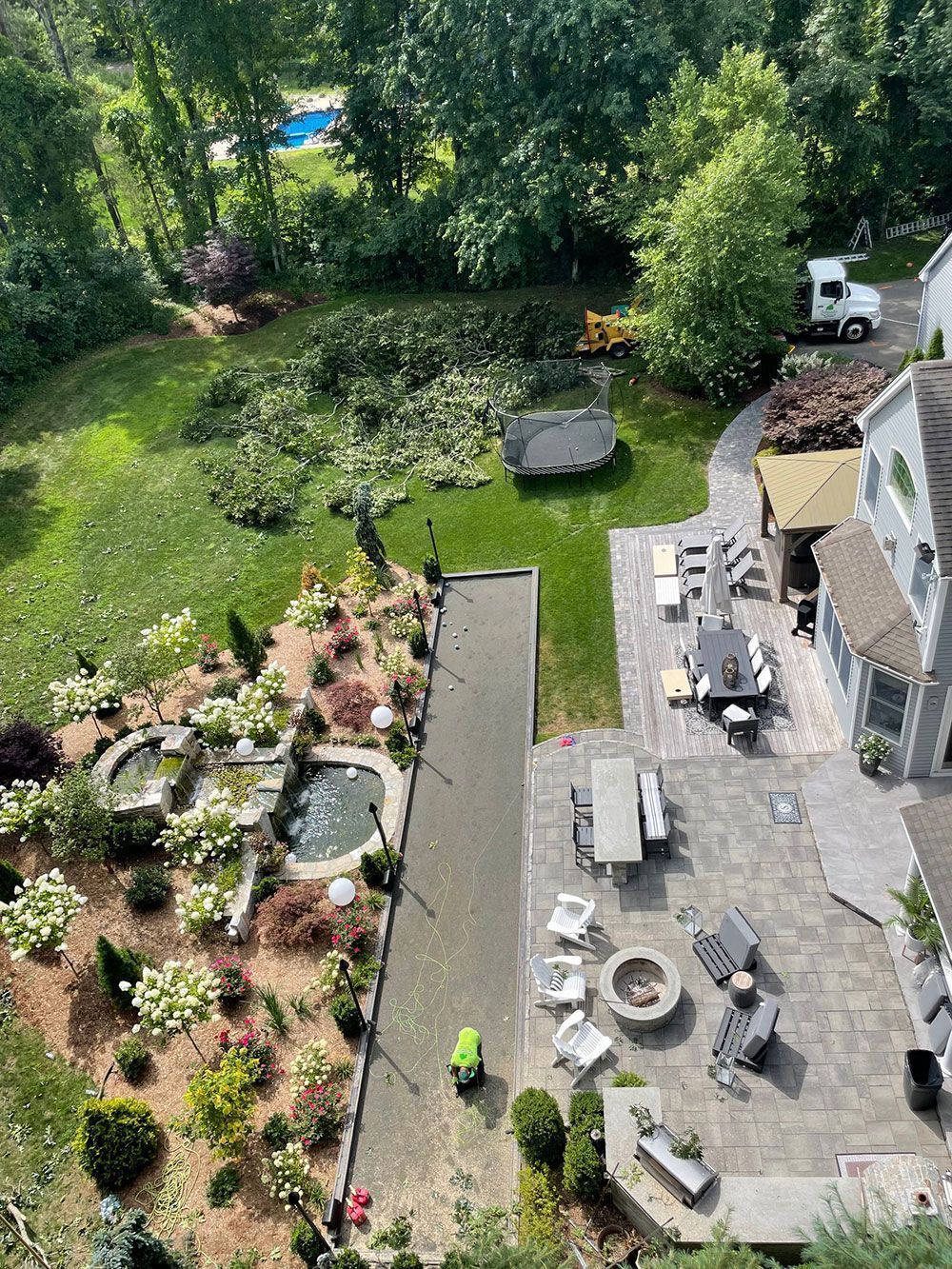 Aerial view of a backyard with garden beds, a narrow pond, patio seating, and a stone house.
