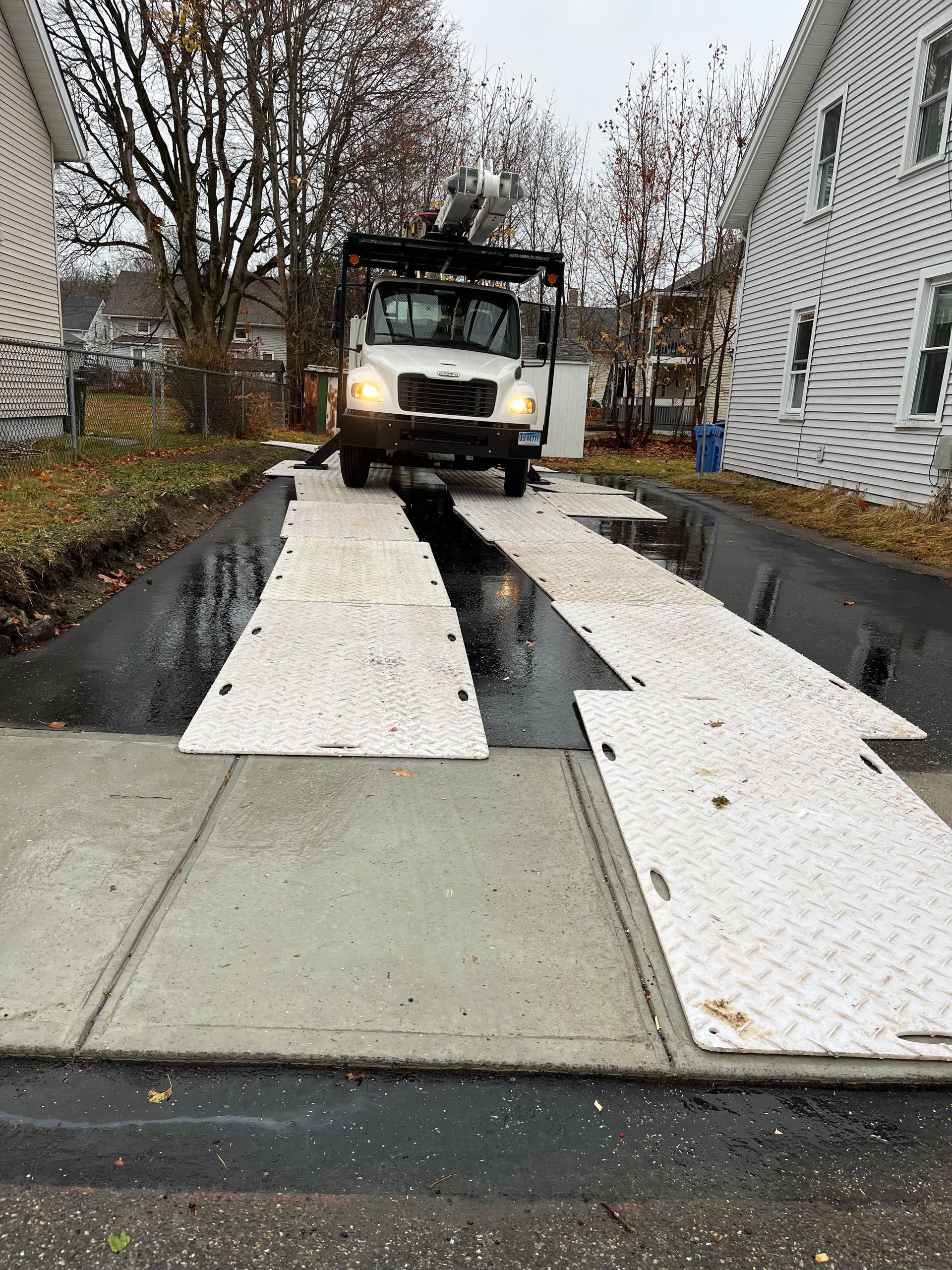 Construction vehicle on a wet driveway between houses, with white planks laid over the pavement.