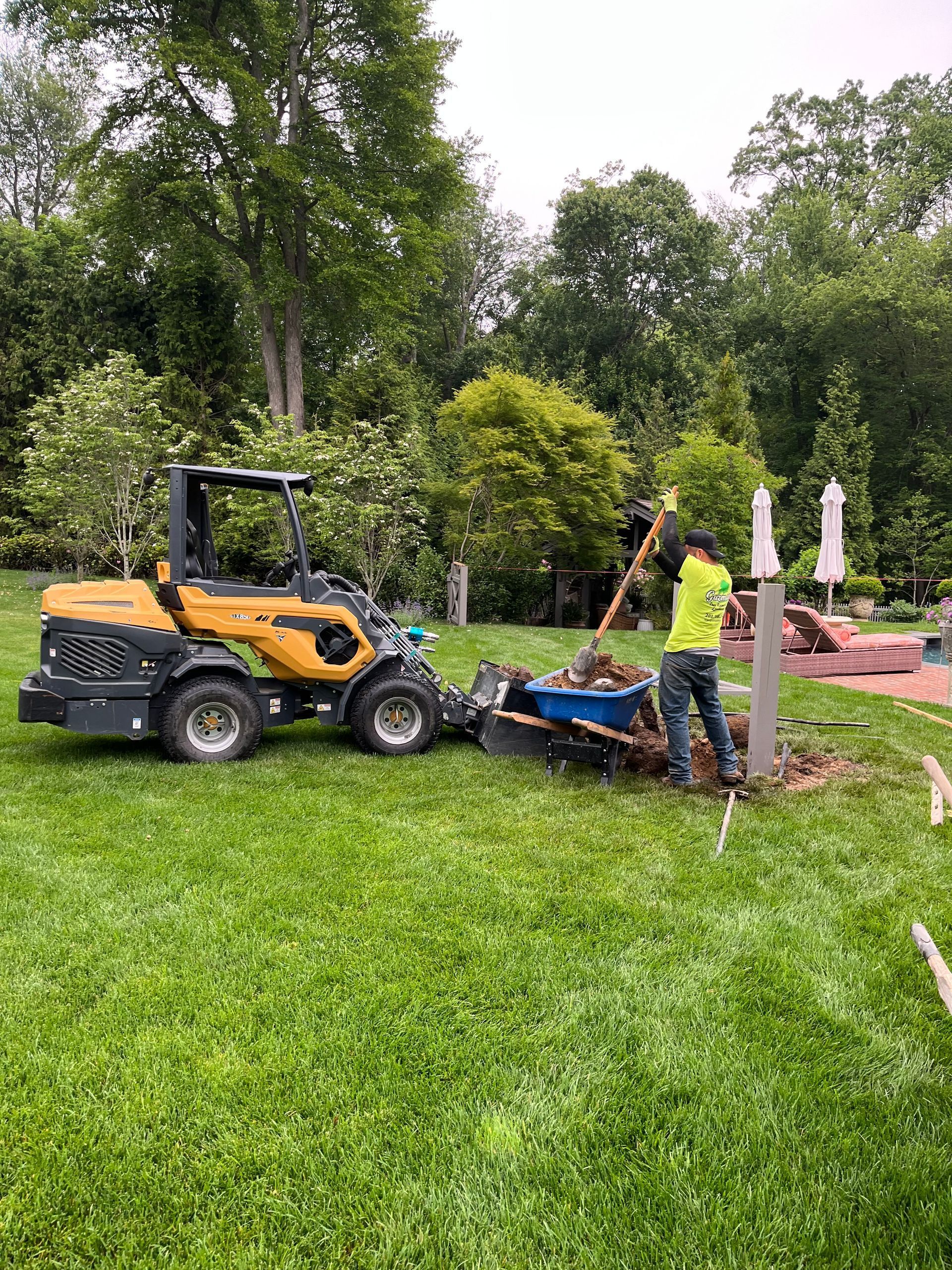Two workers using a compact loader and soil spreader in a grassy garden beside trees and patio umbrellas