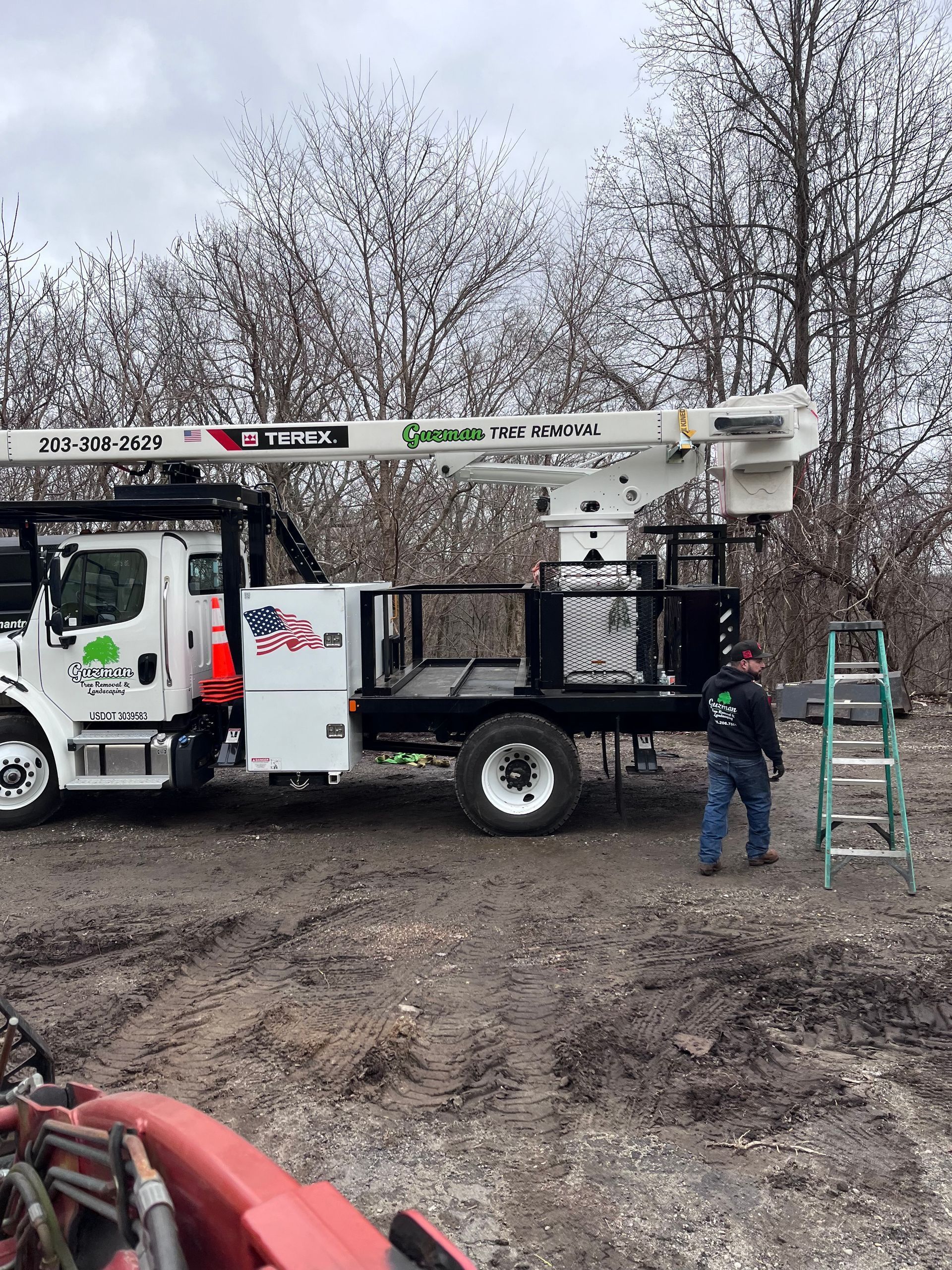 Utility truck with boom lift on muddy roadside, worker standing beside it and a ladder leaning nearby.