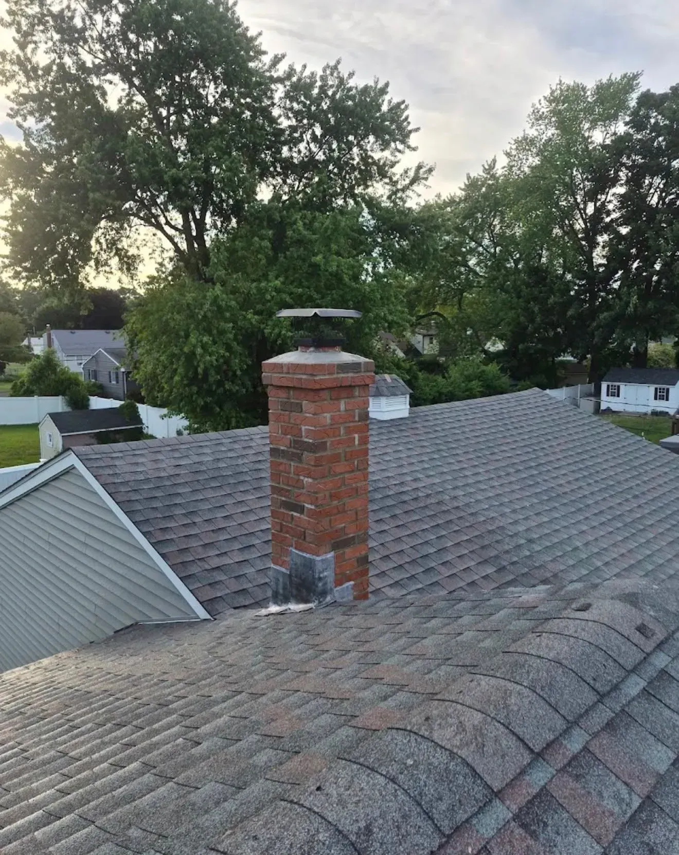 Rooftop with a brick chimney and gray shingles, surrounded by leafy trees at dusk