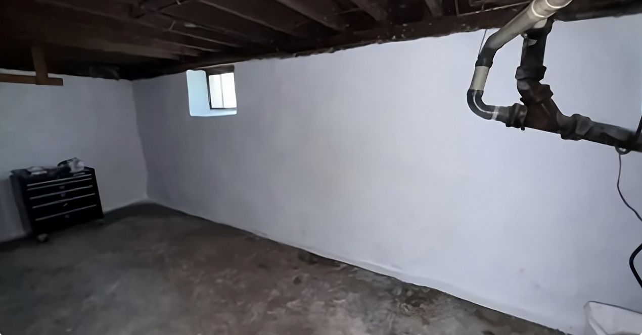 Basement corner with white walls, exposed ceiling pipes, concrete floor, and a small window.
