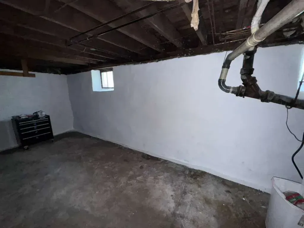 Basement with low exposed wooden ceiling, white walls, small window, and pipes along the right side.